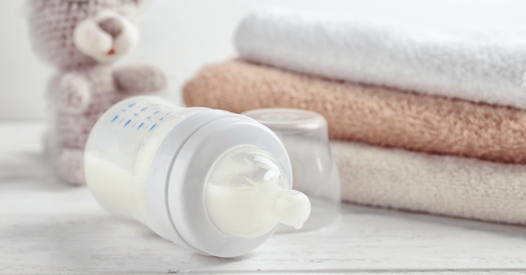 baby bottle on white counter in front of small teddy bear toy and three terry fabric towels in neutral brown and white colours