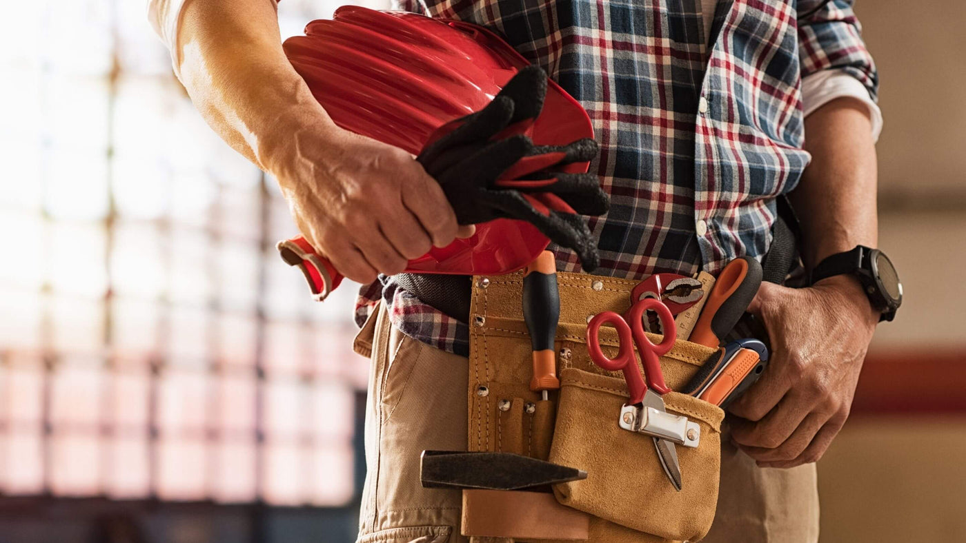 man wearing tool belt filled with a range of tools and holding red hard hat under his arm