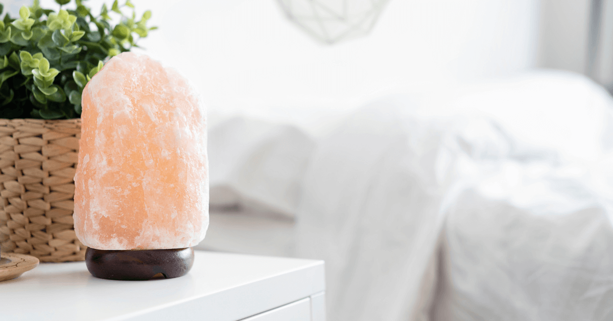 salt lamp on bedroom side table in front of plant in wicker pocket, with bed sheets visible in the background