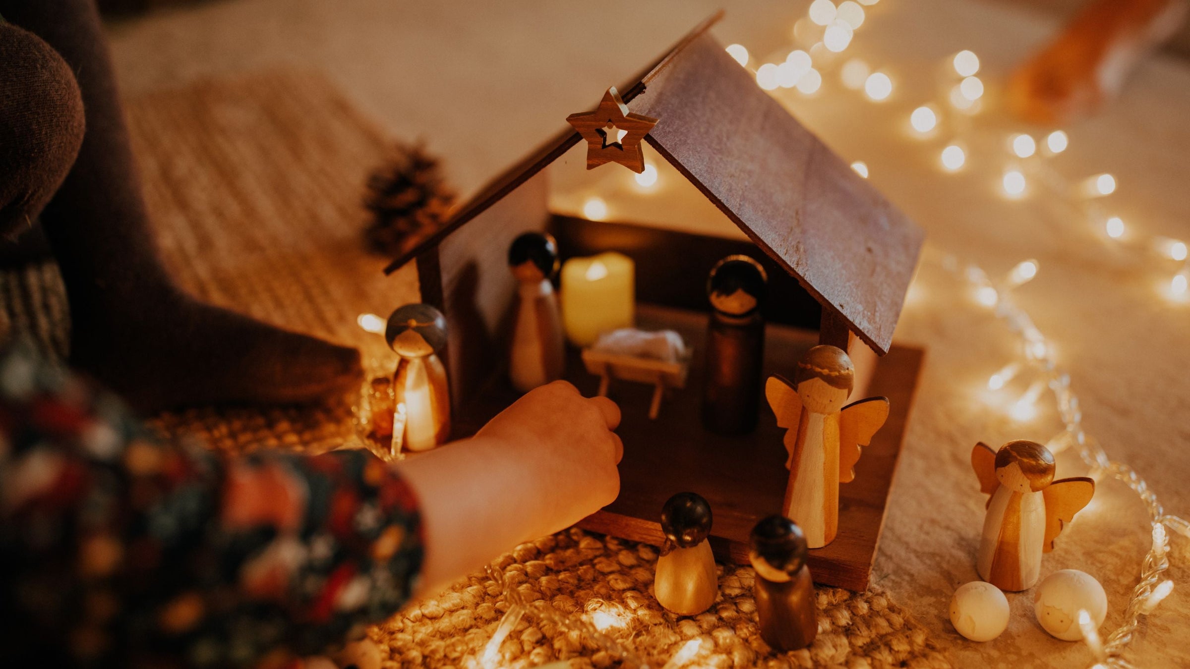 childs hand plays with wooden nativity figures in wooden house on floor with led fairy lights in background