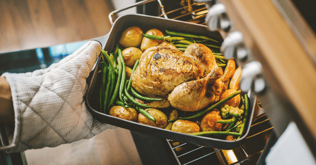 metal oven dish resting on oven shelf with cooked chicken and vegetables inside and oven glove holding the dish