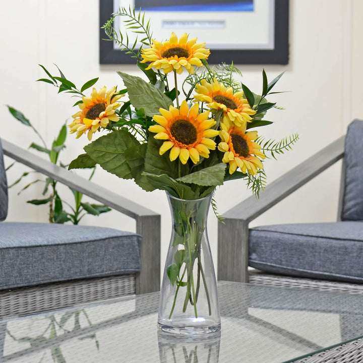Bouquet of sunflowers in a clear vase on a glass table with a chair and plant in the background.