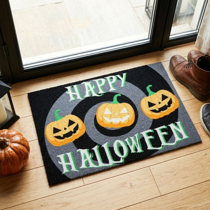 A rectangular Halloween doormat placed on a light wood floor in front of a glass door. The mat is black and gray with the words “HAPPY HALLOWEEN” in white and green, plus three orange jack-o&