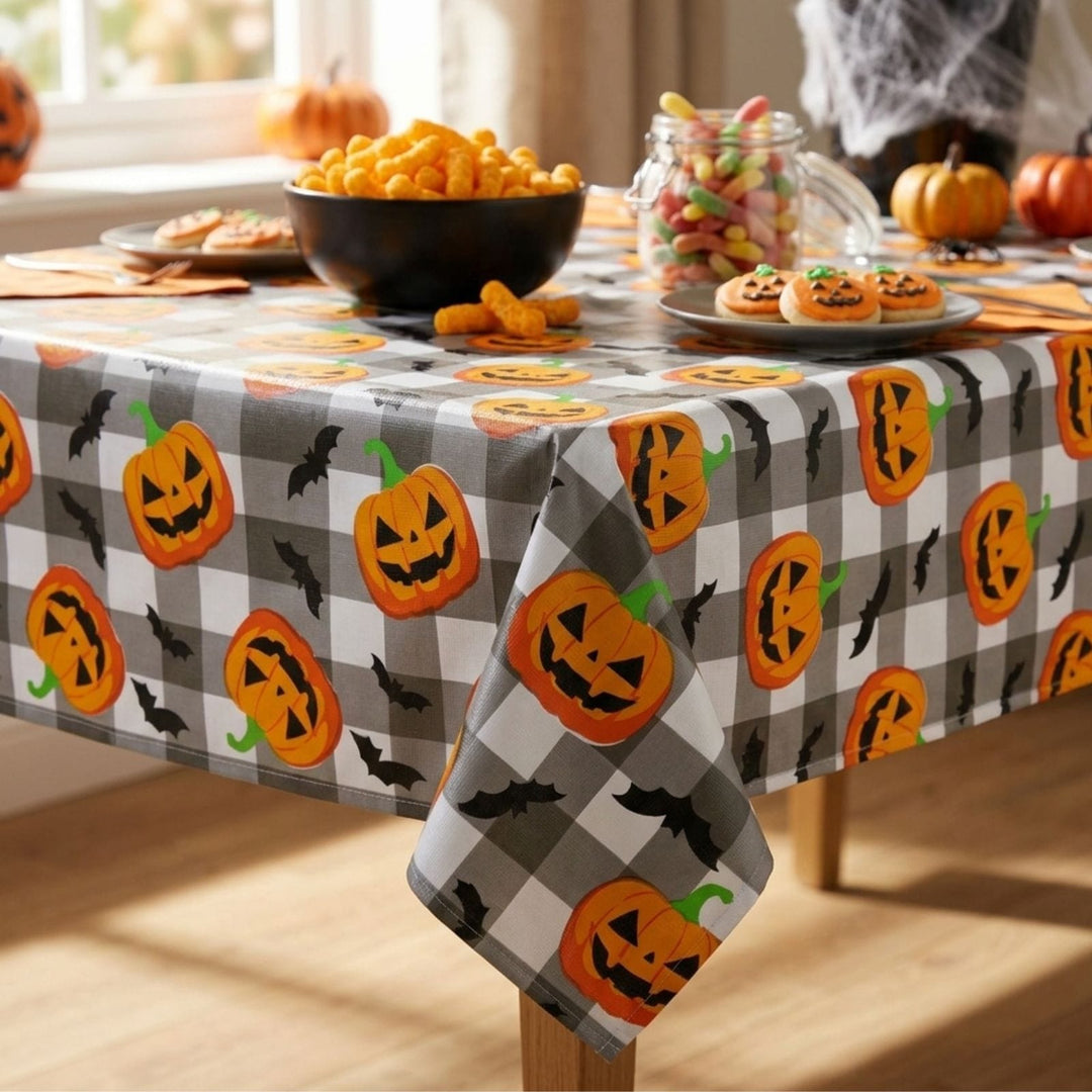 A Halloween-themed tablecloth with a gray and white buffalo check pattern, decorated with orange jack-o'-lanterns and black bats. The tablecloth is shown styled on a wooden table with seasonal snacks, candy in a glass jar, and pumpkins in a brightly lit room.