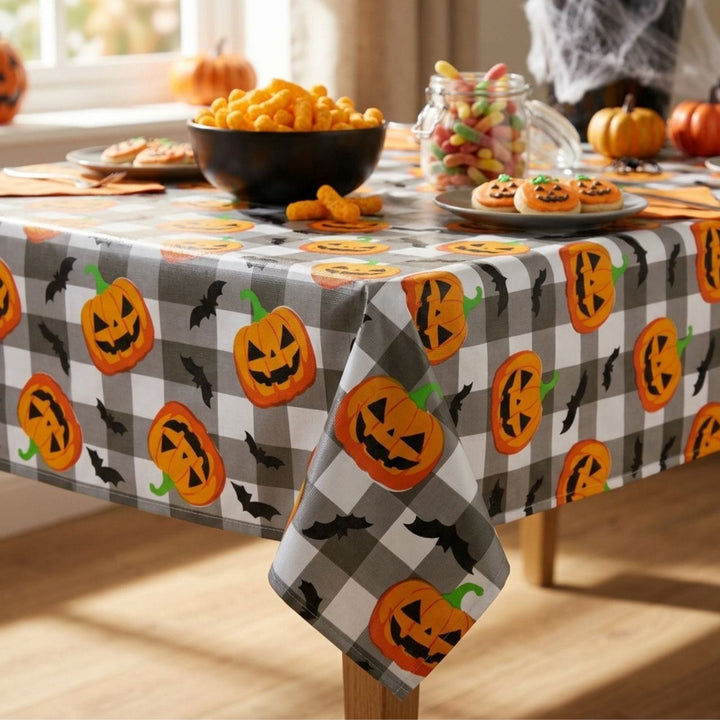 A Halloween-themed tablecloth with a gray and white buffalo check pattern, decorated with orange jack-o'-lanterns and black bats. The tablecloth is shown styled on a wooden table with seasonal snacks, candy in a glass jar, and pumpkins in a brightly lit room.