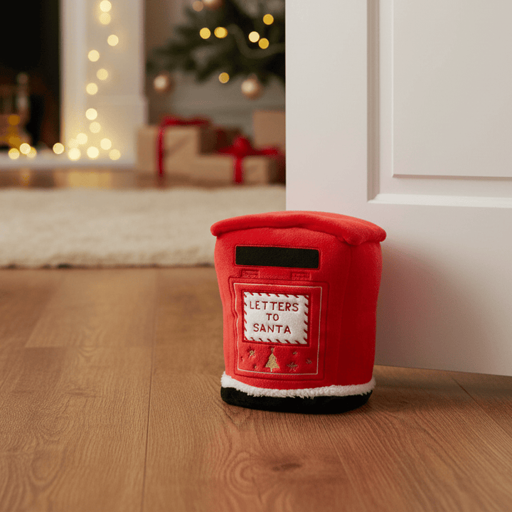 A red plush postman-shaped door stopper with a front patch reading 'LETTERS TO SANTA' sits on a wooden floor in a doorway, with a blurred Christmas tree and fireplace in the background.