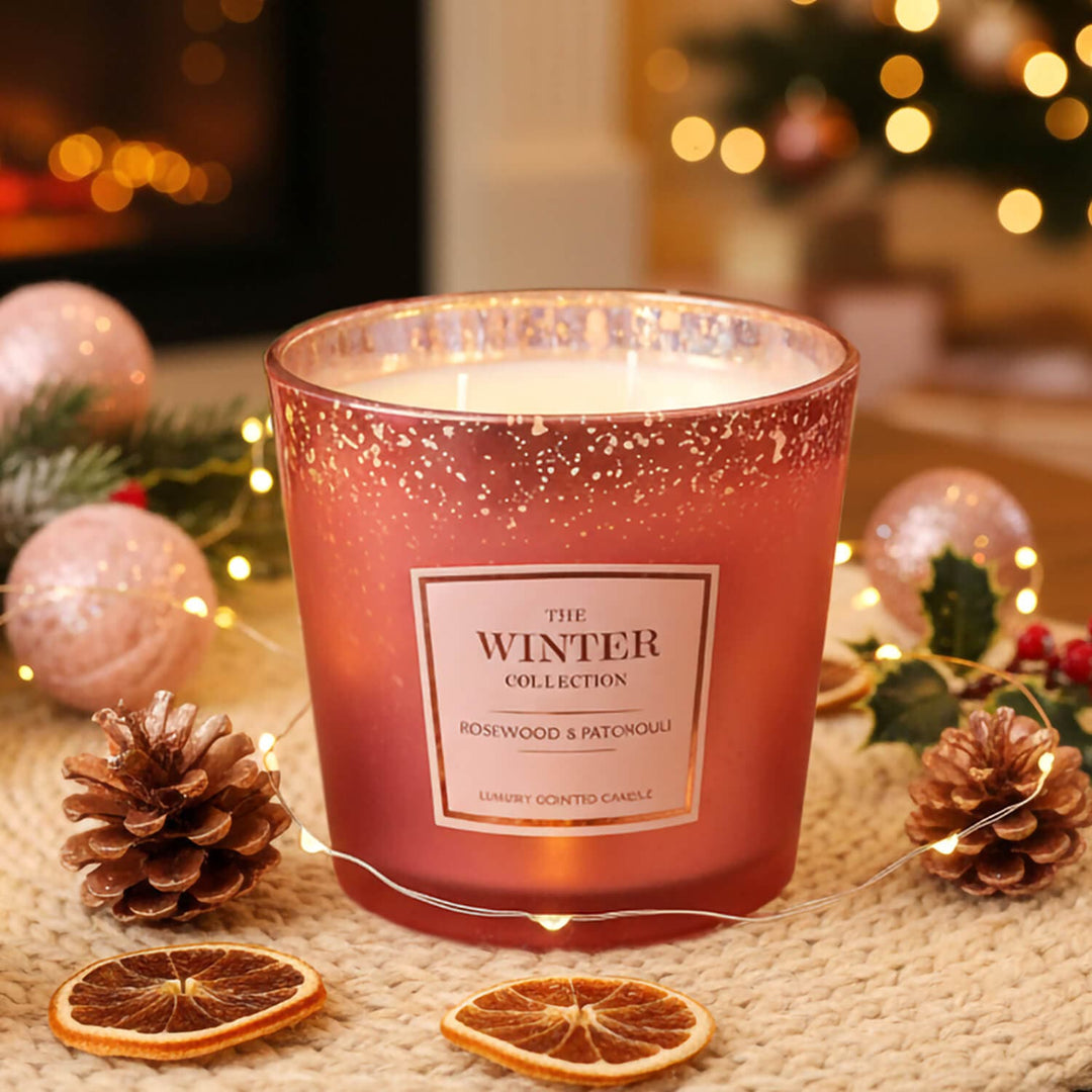A Rosewood &amp; patchouli scented rose gold colour metallic candle jar with a glittery finish and on a table with dried orange slices, pinecones, fairy lights, and holiday decorations in the background.