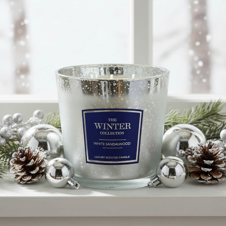 A white sandalwood scented candle in a silver glittery glass jar, styled on a white windowsill with silver ornaments, pinecones, and green pine branches. 
