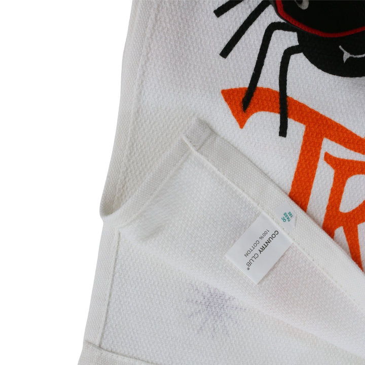 Close-up of a white textured fabric garment with a black spider graphic, orange lettering, and a sewn-in size tag reading &