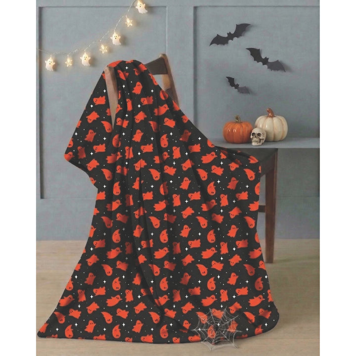 A black blanket or throw draped over a wooden chair in a styled room, featuring an all-over orange bat pattern with small white stars. The background includes a gray paneled wall, hanging string lights, black bat cutouts, a small pumpkin, a skull, and a spiderweb decoration.
