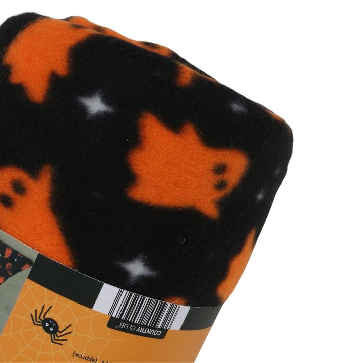 A close-up of a folded fleece blanket with a Halloween pattern featuring orange ghosts and white stars on a black background, with a cardboard band showing a barcode and spider graphic.