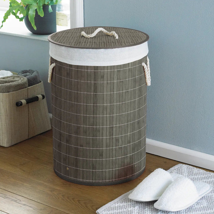 Grey and white bamboo laundry basket with white rope handles in a room with slippers and towels