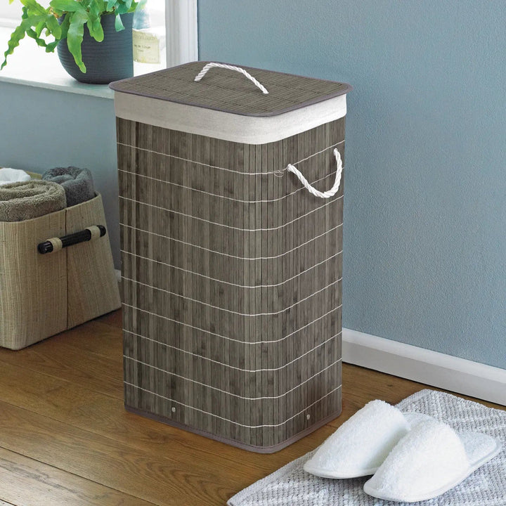 Grey bamboo and rattan laundry hamper with white rope handles in a room with slippers and towels