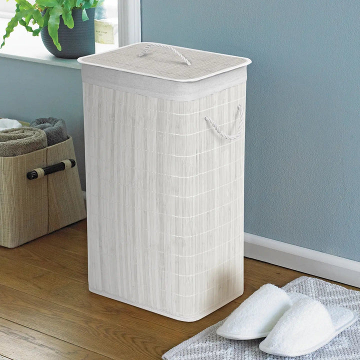 White bamboo and rattan laundry hamper with white rope handles in a room with slippers and towels