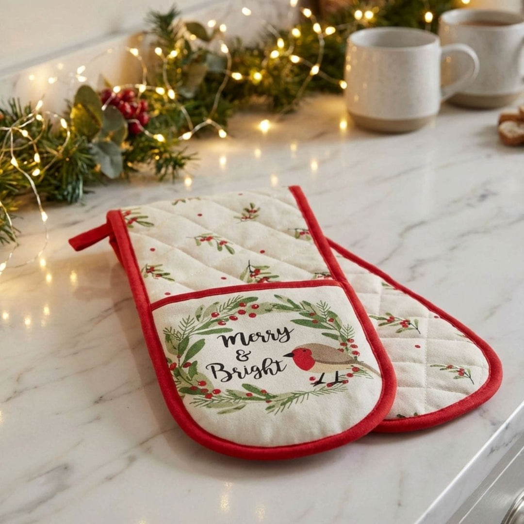 Pair of Christmas-themed oven mitts on a marble countertop with decorative lights and mugs in the background.