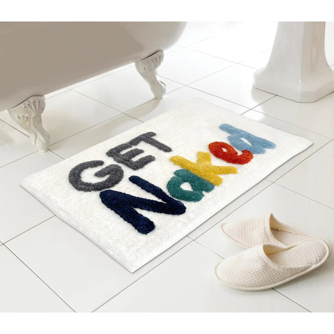 Bath mat with Get Naked letters in bright colours on a tiled floor next to a bath and slippers