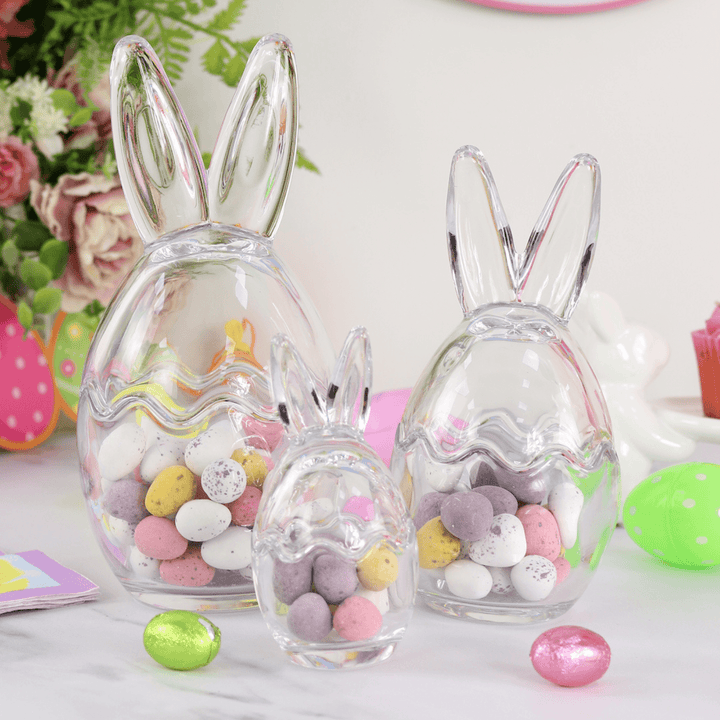 Set of three glass jars with removable bunny ear lids, filled with various colorful candies, displayed on a table with Easter decorations.