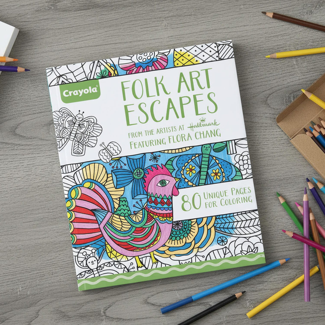 A Crayola Folk Art Escapes coloring book is displayed on a gray wood-textured surface, surrounded by colored pencils and a pencil box.