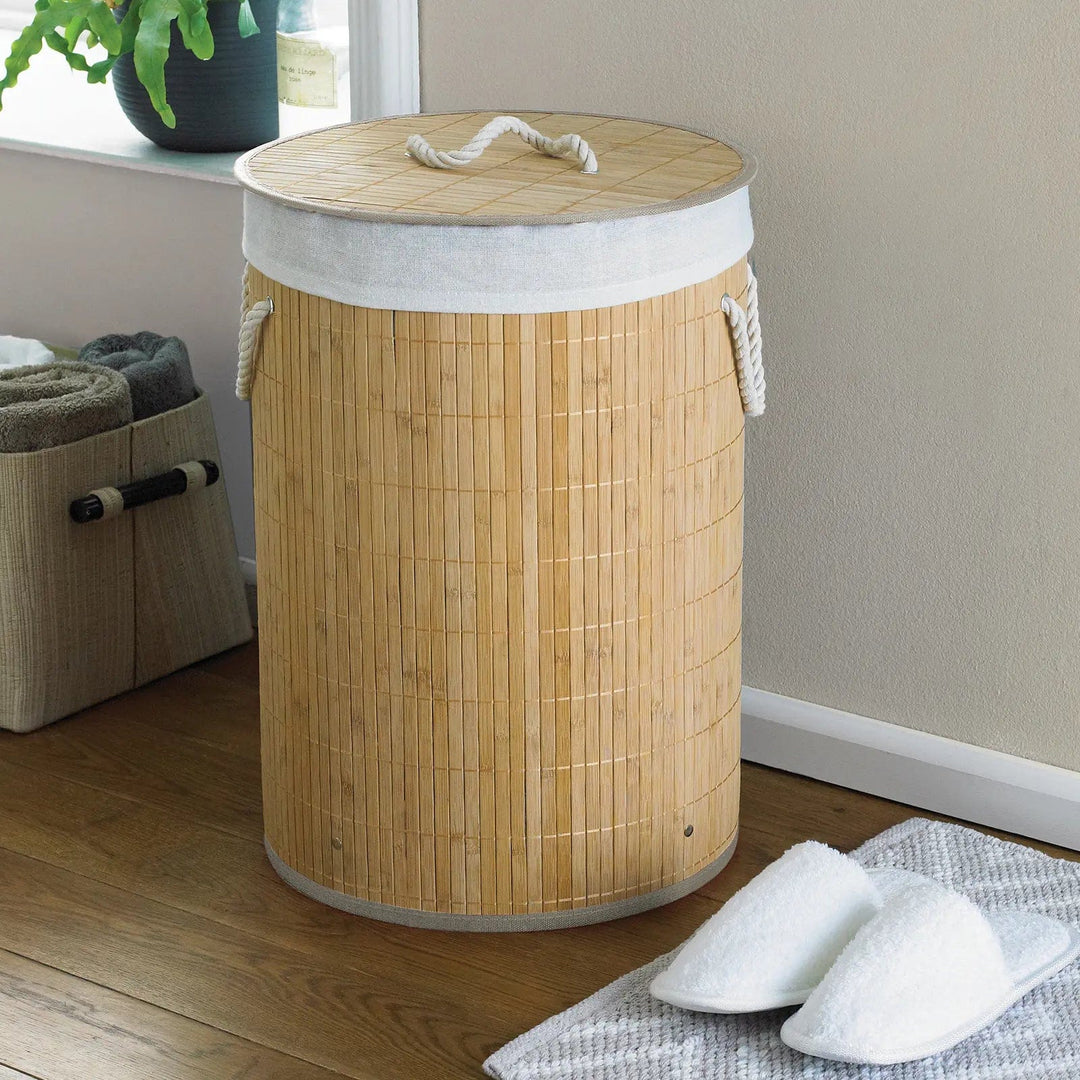 Round natural colour bamboo laundry basket with rope handles in a bathroom with white slippers and towel bag