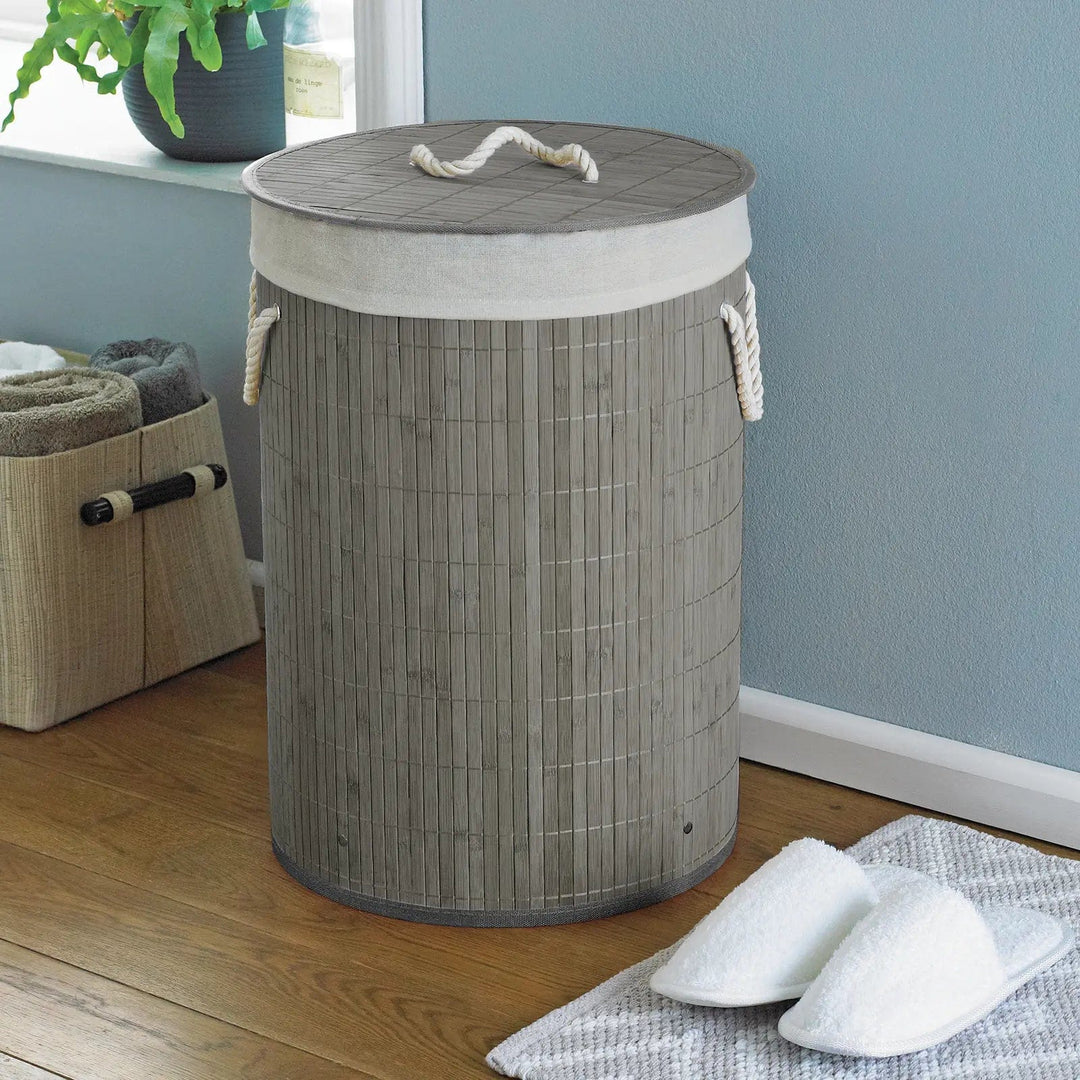 Warm grey natural bamboo laundry basket with lid and rope handles, in a bedroom with slippers and towel holder