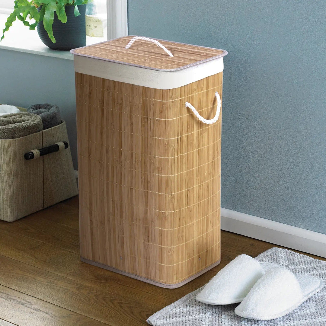 Natural colour bamboo laundry basket with white rope handles, in a bathroom with slippers and towel storage
