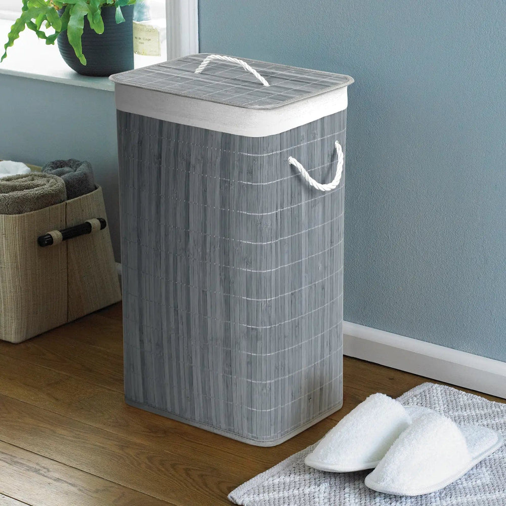 Grey, natural bamboo laundry basket with white rope handles, in a bathroom with slippers and towel storage