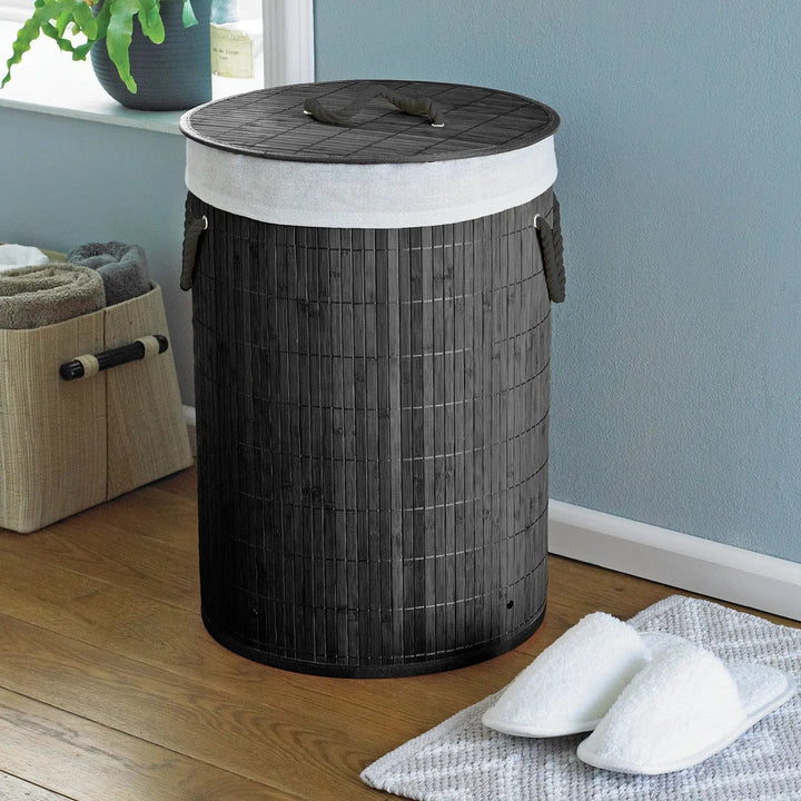 Charcoal grey natural bamboo laundry basket with lid and rope handles, in a bedroom with slippers and towel holder