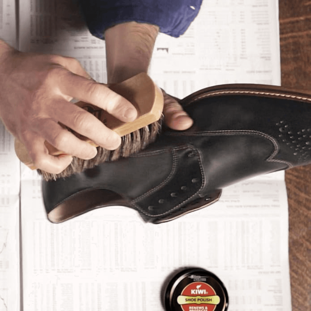 person buffing a black shoe with brush and kiwi polish in background