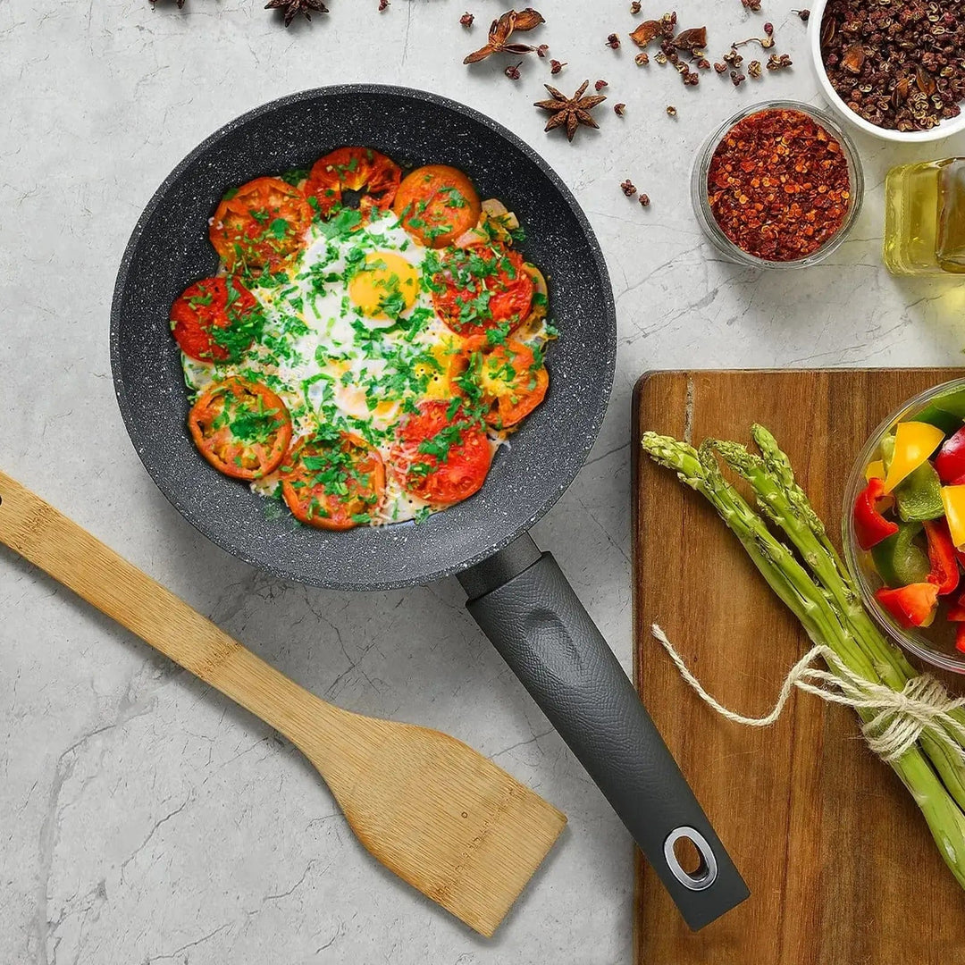 Overhead view of a black frying pan filled with tomato omelette on a worktop with chopping board, asparagus, chopped peppers, wooden spoon, herbs and spices