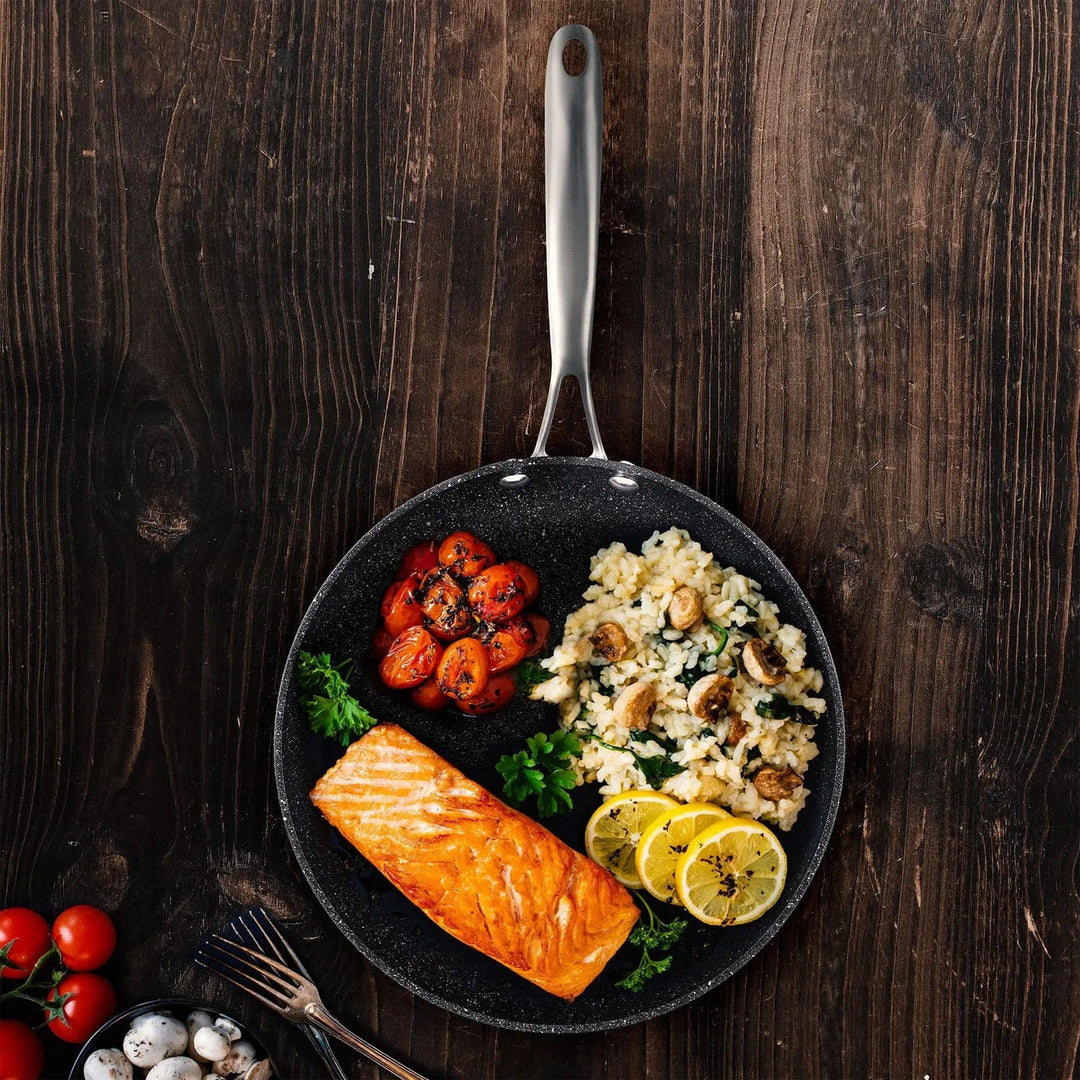 Overhead view of a large black frying pan filled with salmon fillet, tomatoes, lemon silces and mushroom risotto, on a dark wooden table