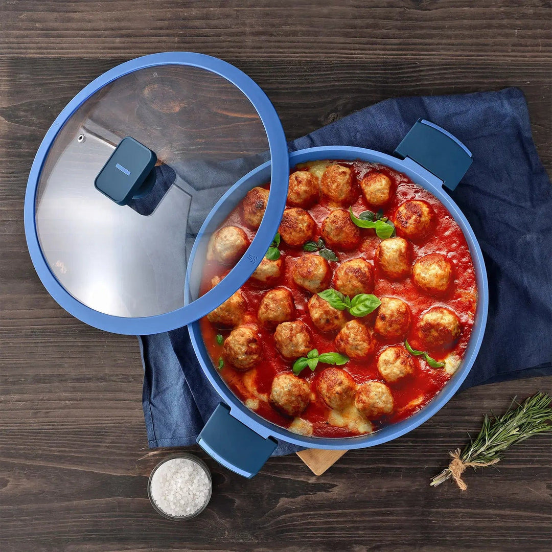 Casserole dish in blue with glass lid, filled with meatballs in tomato sauce, on a wooden table with salt and herbs