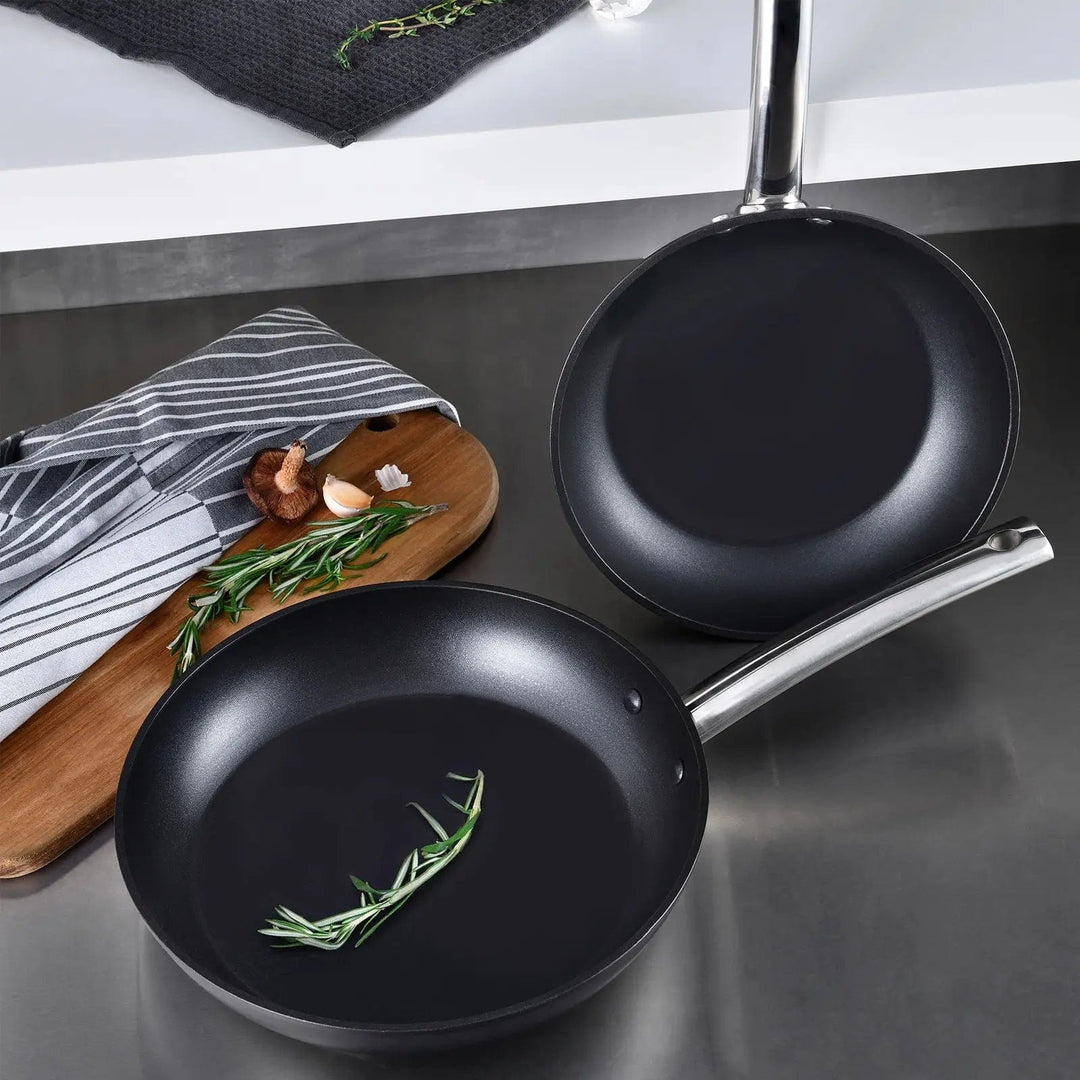 2 Foodies range frying pans with matt black finish and silver handles on a worktop with chopping board, apron and herbs