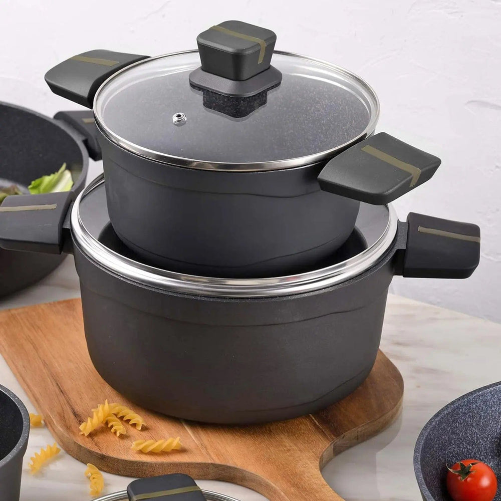 2 black casserole dishes with glass lids and inset handles on a wooden chopping board with pasta and tomatoes