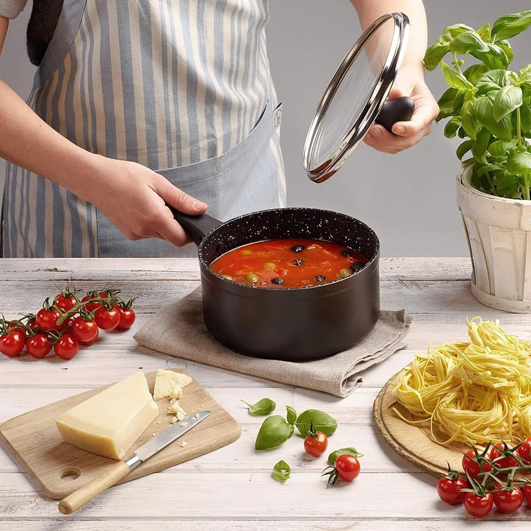 Cook holding a black saucepan filled with tomato sauce and olives, with glass lid, on a table with tomatoes, basil plant, parmesan cheese and pasta