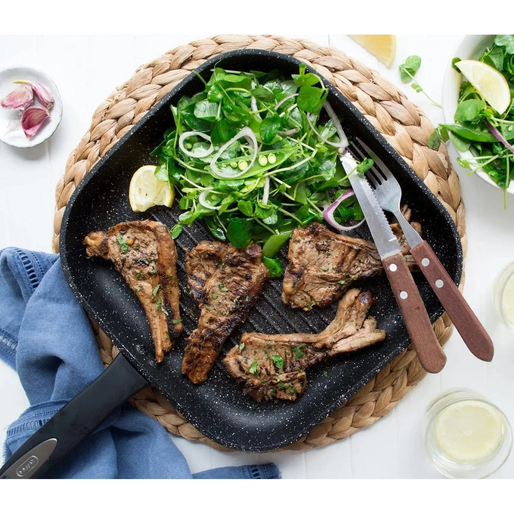 Black square grill pan filled with grilled chops, salad leaves, knife and fork, on a woven mat, with garlic and dips