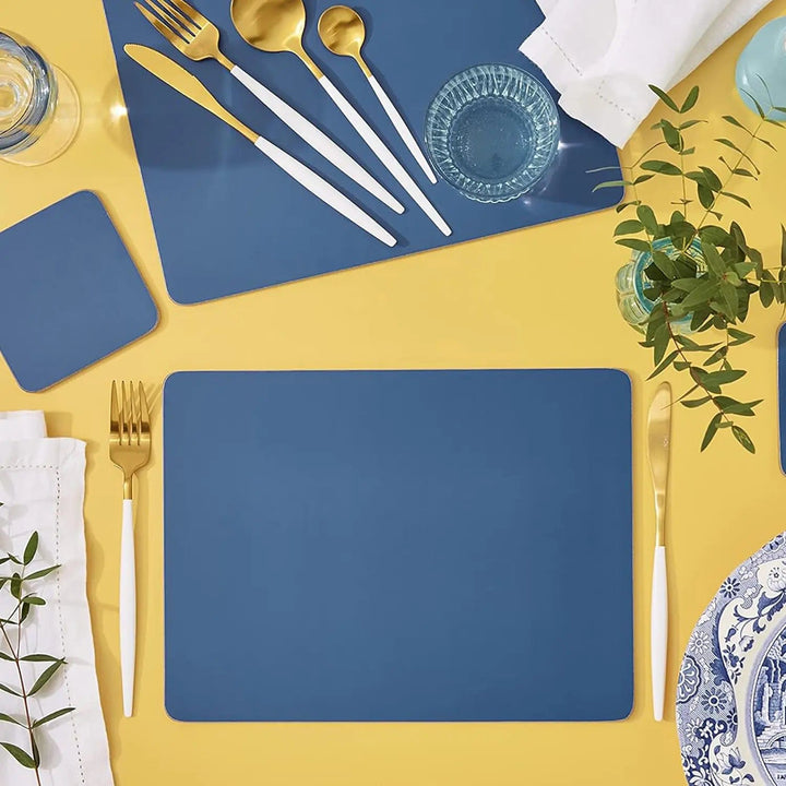 Blue placemats and coaster on a yellow table with cutlery, glasses and foliage