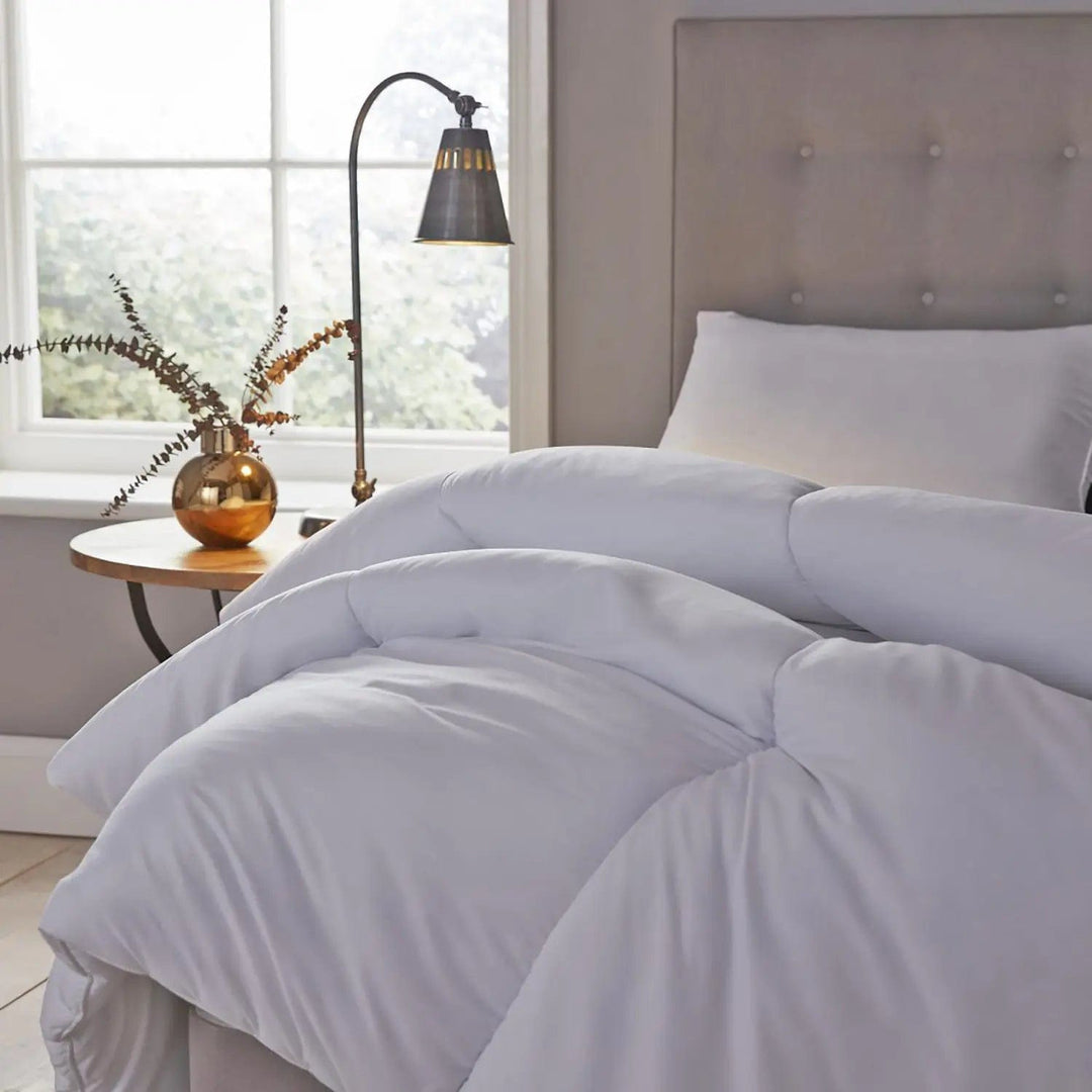 Duvet folded back on a bed with silver headboard, gold vase abd foliage at a window