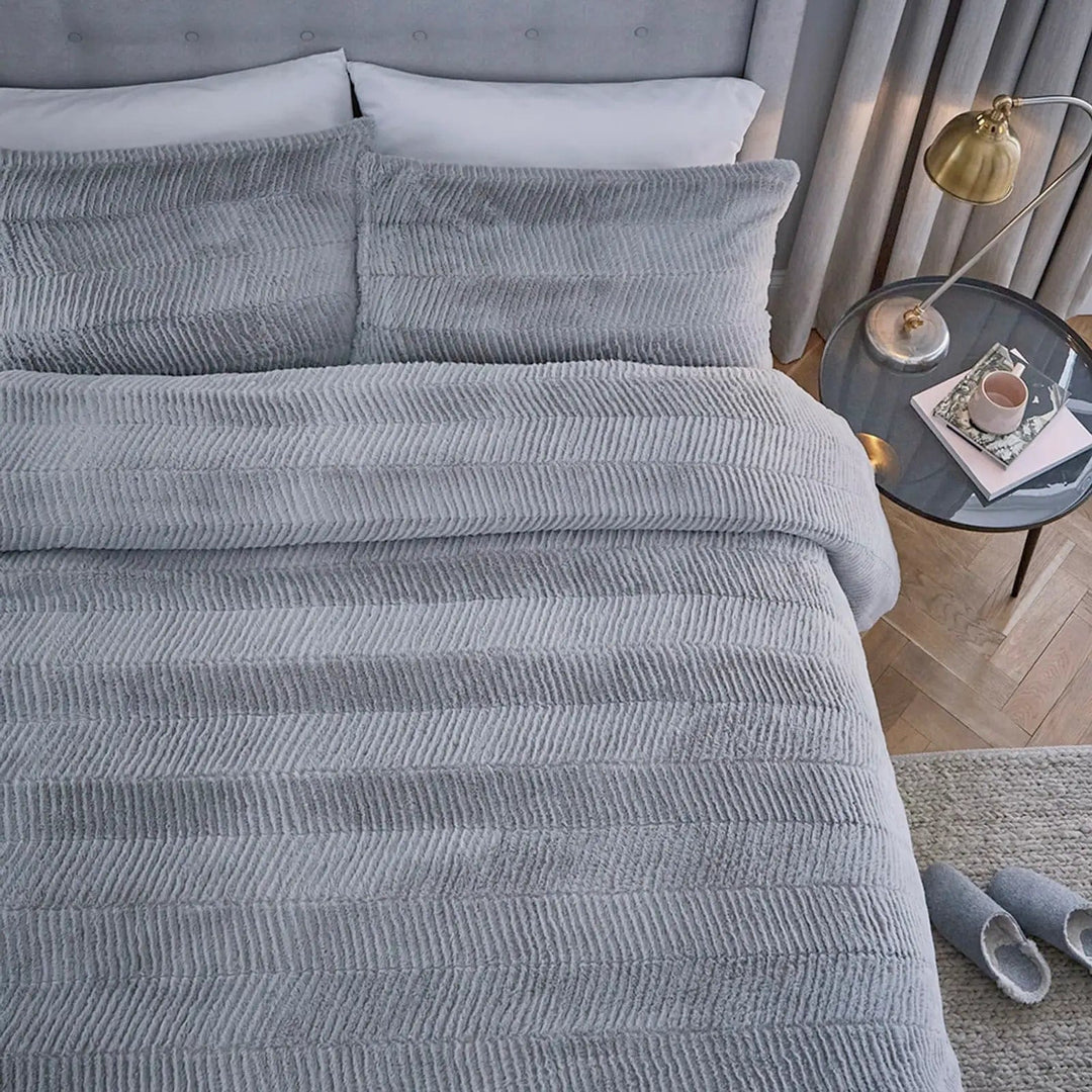 Top view of grey chevron pattern faux fur quilt cover and pillowcases on a bed with side table, rug and slippers