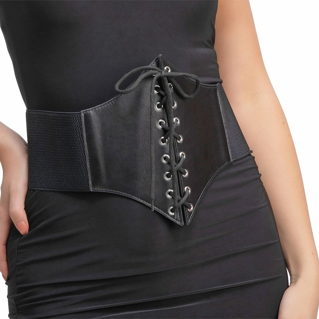 A close-up of a model wearing a black waist corset belt with front lace-up detailing and silver eyelets, paired with a black dress against a white background.