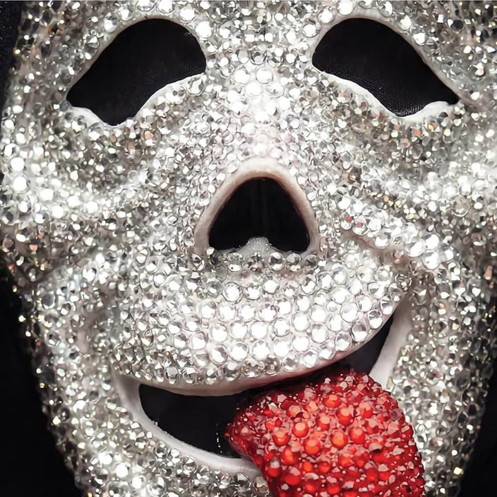 A close-up of a silver rhinestone mask with a red rhinestone tongue sticking out against a black background.