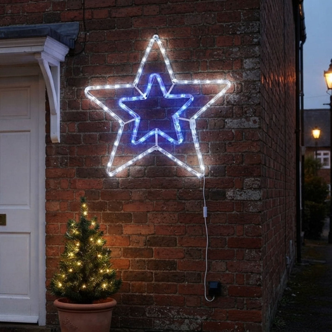 A large five-pointed star LED light decoration mounted on a red brick wall, with a glowing blue inner star and white outer star. A small potted Christmas tree with warm lights sits below on the left, and a house corner and street scene are visible in the background at dusk.
