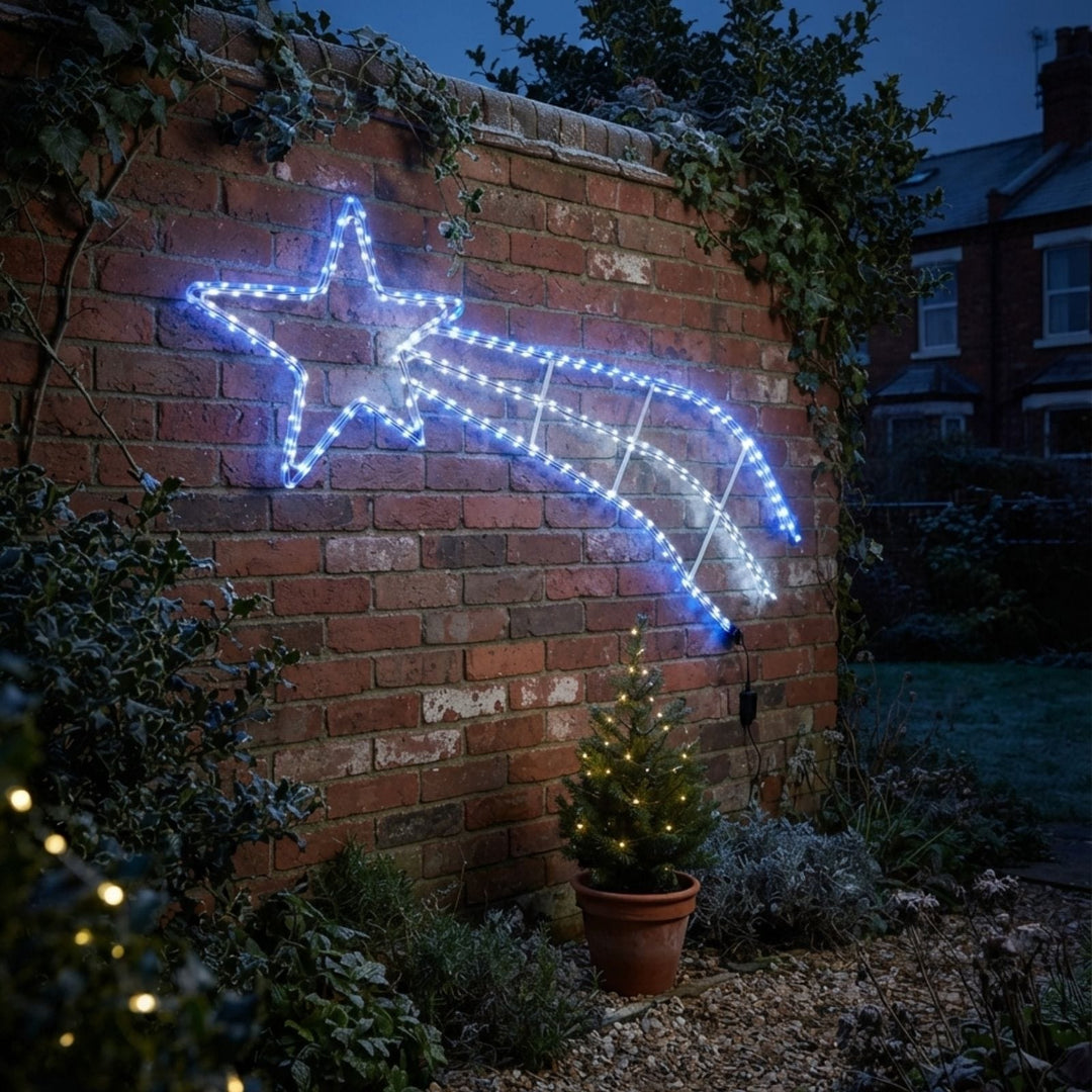 A glowing star-shaped light with a shooting-stripe effect mounted on a red brick wall outdoors at dusk, with plants, a small potted tree, and a house visible in the background.