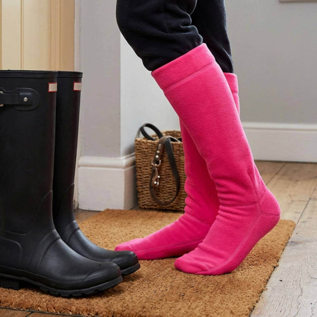 A person wearing bright pink knee-high wellington boot liners standing on a brown door mat next to a pair of black rain boots in an indoor setting.
