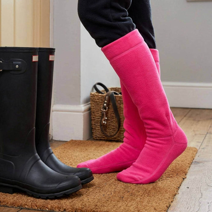 A person wearing bright pink knee-high wellington boot liners standing on a brown door mat next to a pair of black rain boots in an indoor setting.