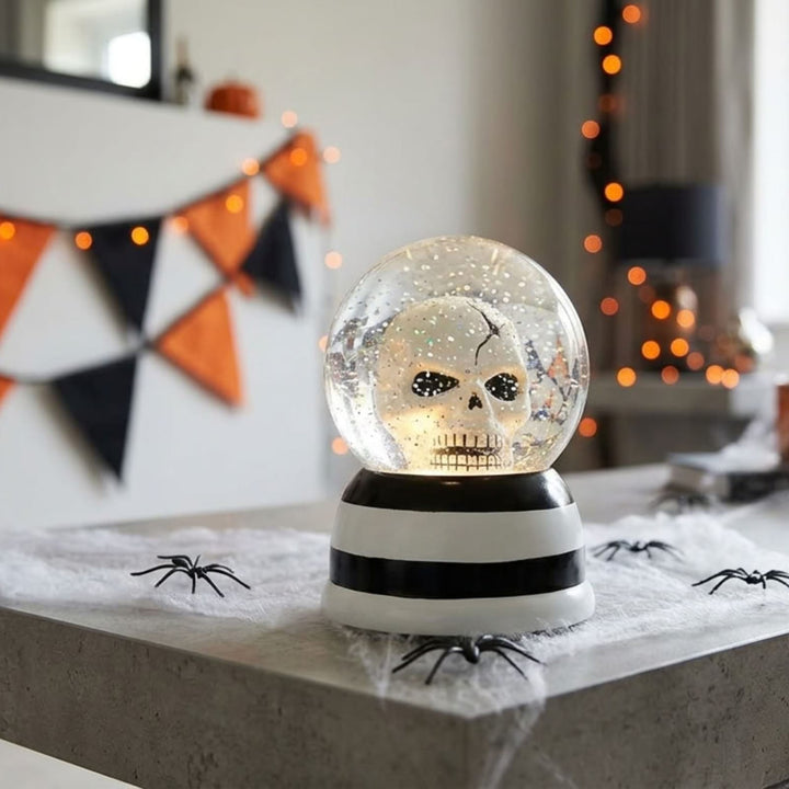 A Halloween-themed snow globe with a skull design inside, sitting on a gray stone surface decorated with fake spiderwebs and black spiders. The background shows a softly blurred indoor setting with orange bokeh lights and a black, white, and orange triangular banner.