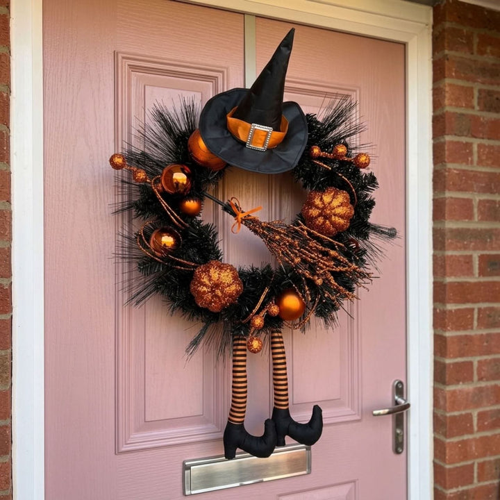 A Halloween witch-themed wreath hanging on a pink front door, decorated with a black pointed hat, orange and gold ornaments, a broom, and black-and-orange striped legs.