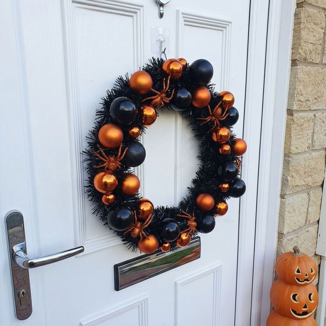 A black Halloween wreath decorated with shiny black and orange baubles, orange spider decorations, and small orange pumpkins, hanging on a white front door.