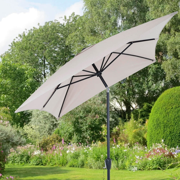Beige parasol, tilted with crank handle in a garden setting with trees and flowers.