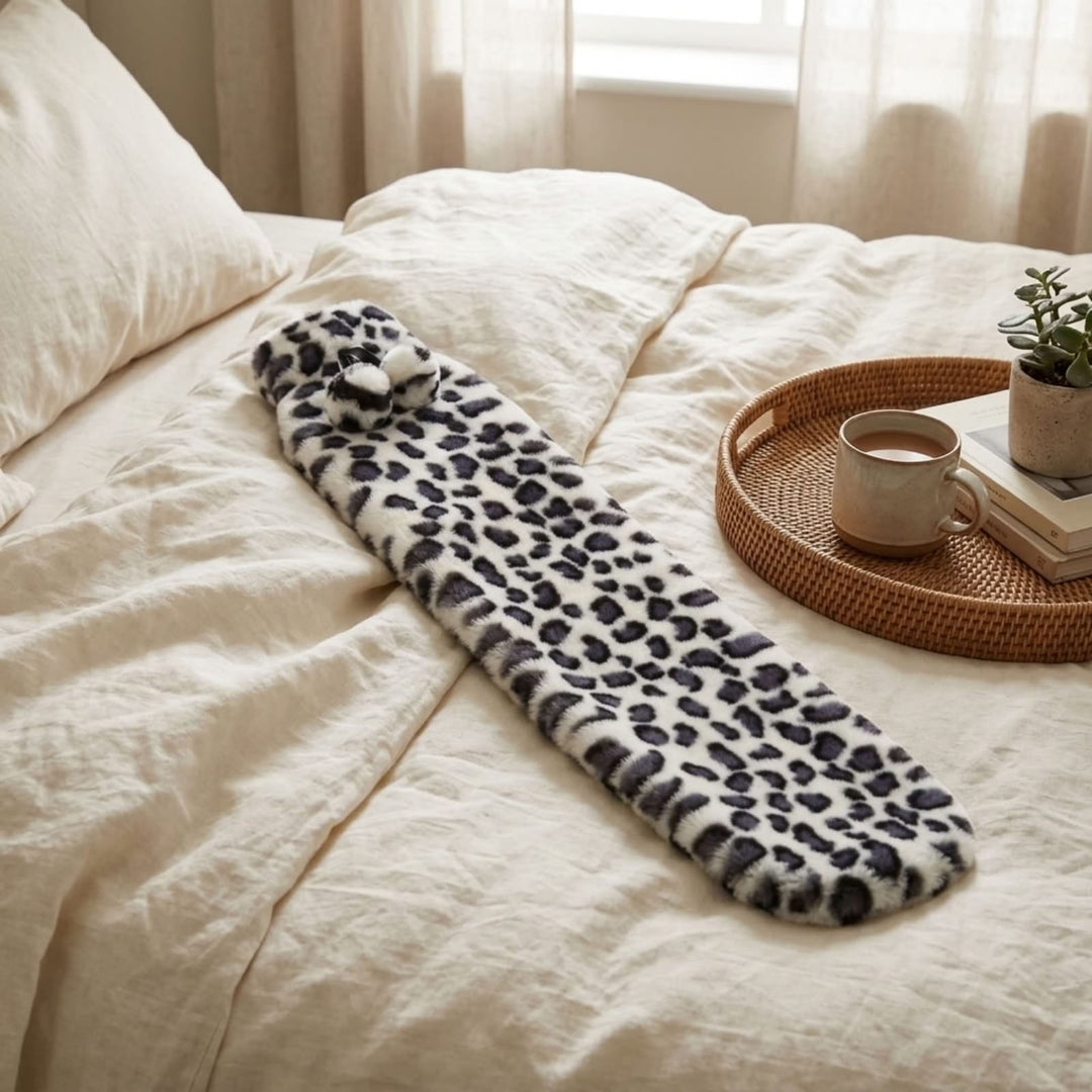A plush fleece hot water bootle with a black and white leopard print pattern is draped on a bed with cream-colored bedding. A woven tray with a mug and a small plant sits nearby on the bed.