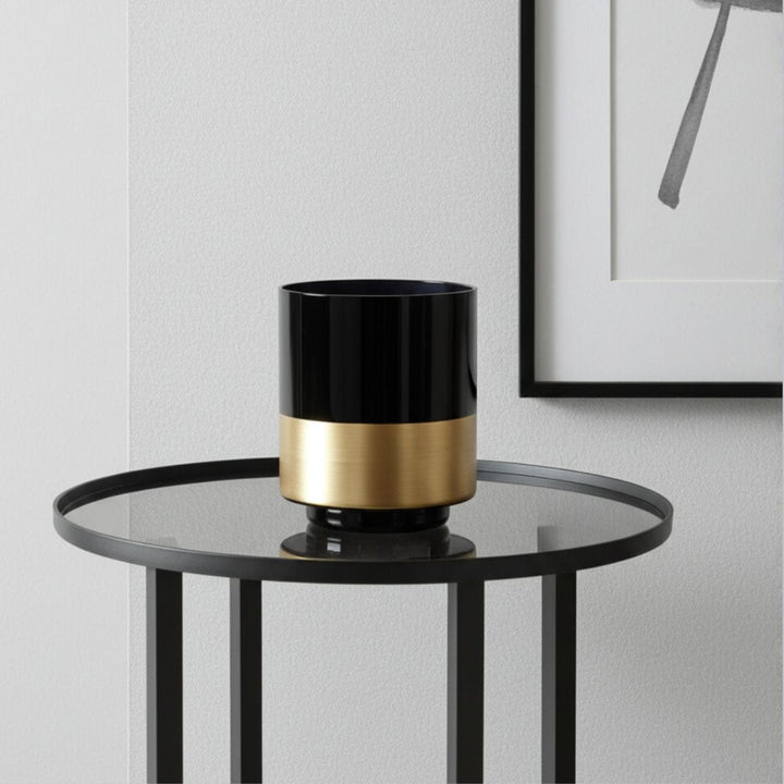 Black and gold cylindrical vase on a round glass table against a light gray wall.